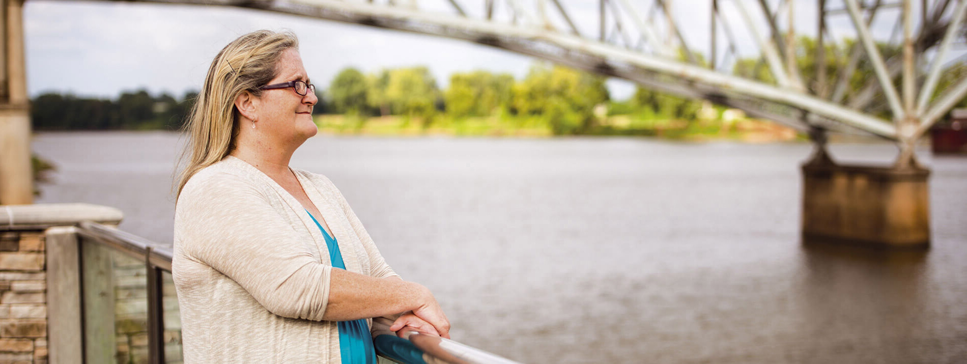 Μία γυναίκα που στέκεται δίπλα σε μία γέφυρα κοιτάζοντας μακριά, πάνω από ένα ποτάμι.