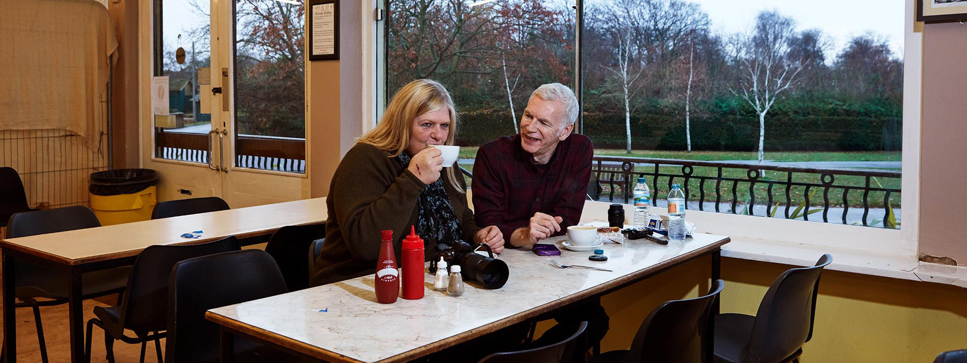 Ένας χαμογελαστός άντρας που κάθεται σε ένα τραπέζι με μια γυναίκα που πίνει ένα ρόφημα από μία κούπα.