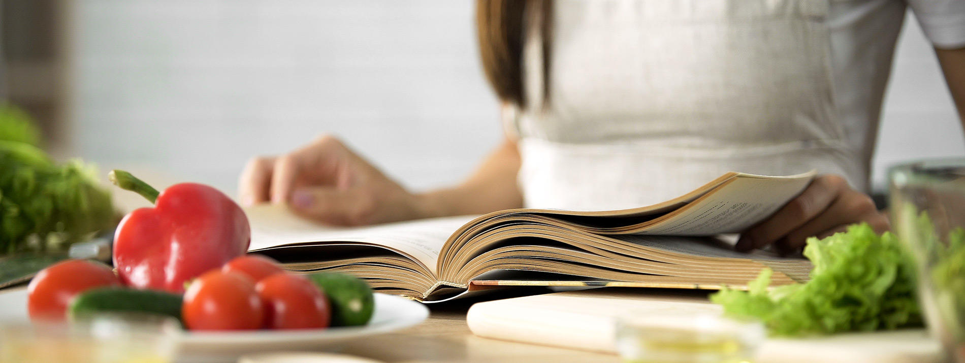 Woman browser through a recipe book with vegatables laying around the table.