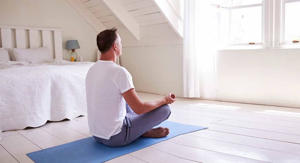 A light room with a man sitting on a blue mat doing yoga.